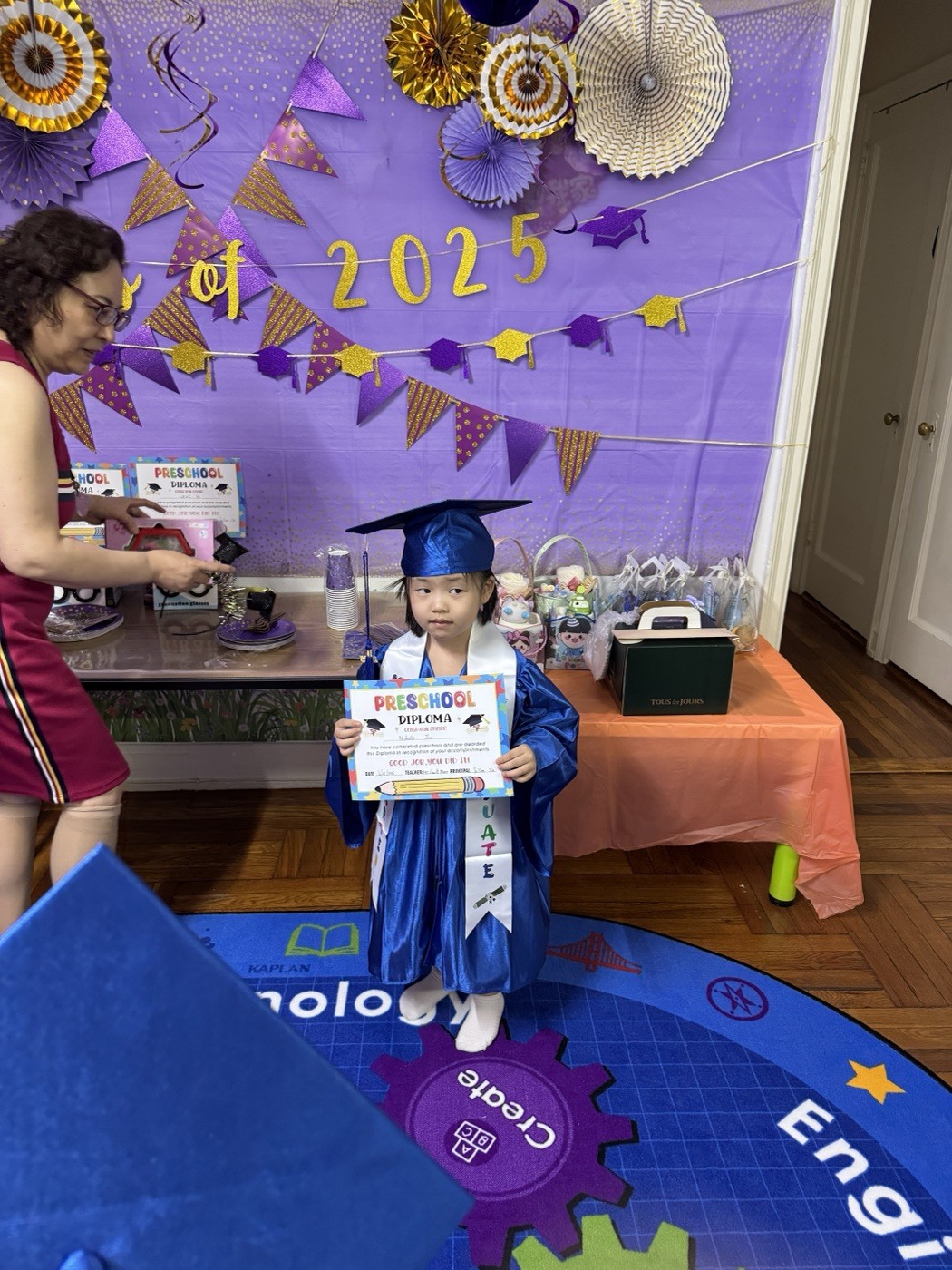 小女孩身穿蓝色学士服手持新乐幼儿园毕业证书站在2025毕业背景墙前