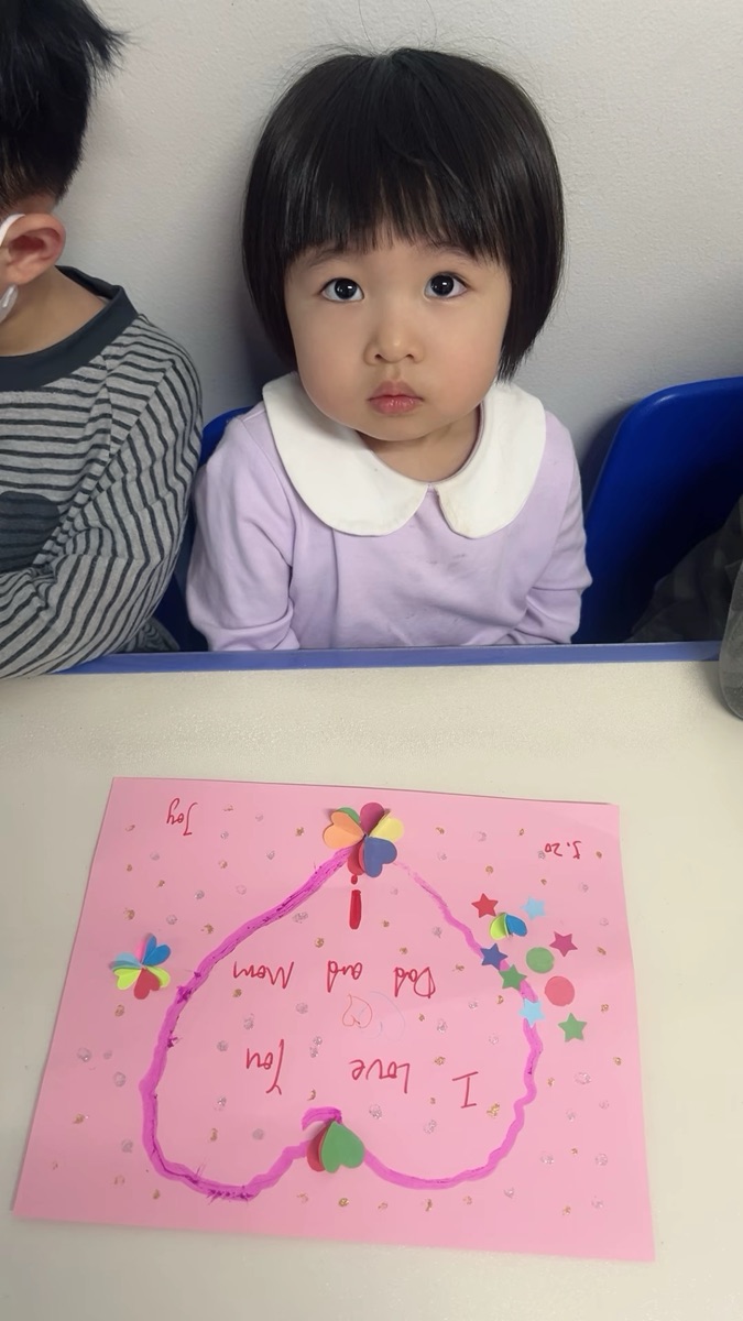小女孩端坐桌前,展示亲手制作的I Love You Dad and Mom粉色爱心贺卡